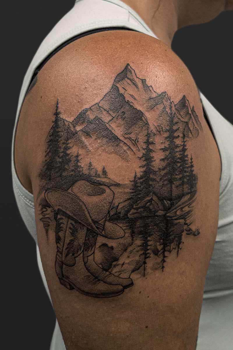 Legacy Tattoo| Black and grey, realism tattoo of a mountain range, forest, cowboy boots and hat