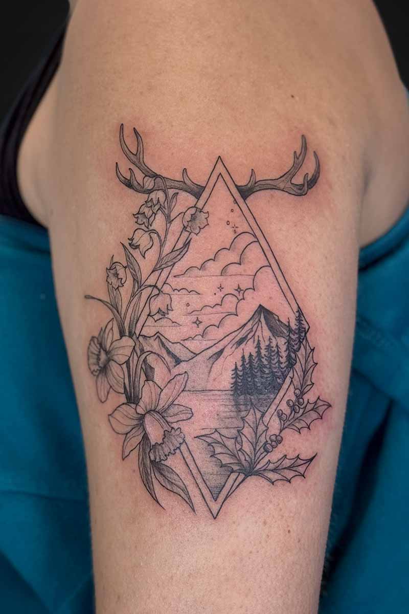 Legacy Tattoo| Black and grey tattoo of a mountain scene, framed in a diamond with florals and antlers