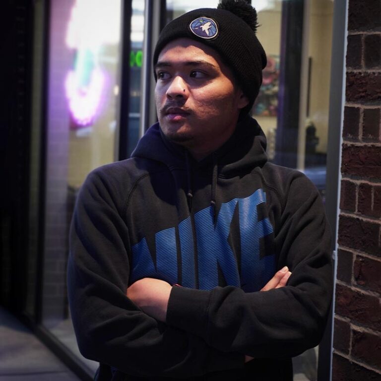 Man in black Nike hoodie and toque with pom-pom standing with arms crossed outdoors at night, neon lights blur behind.