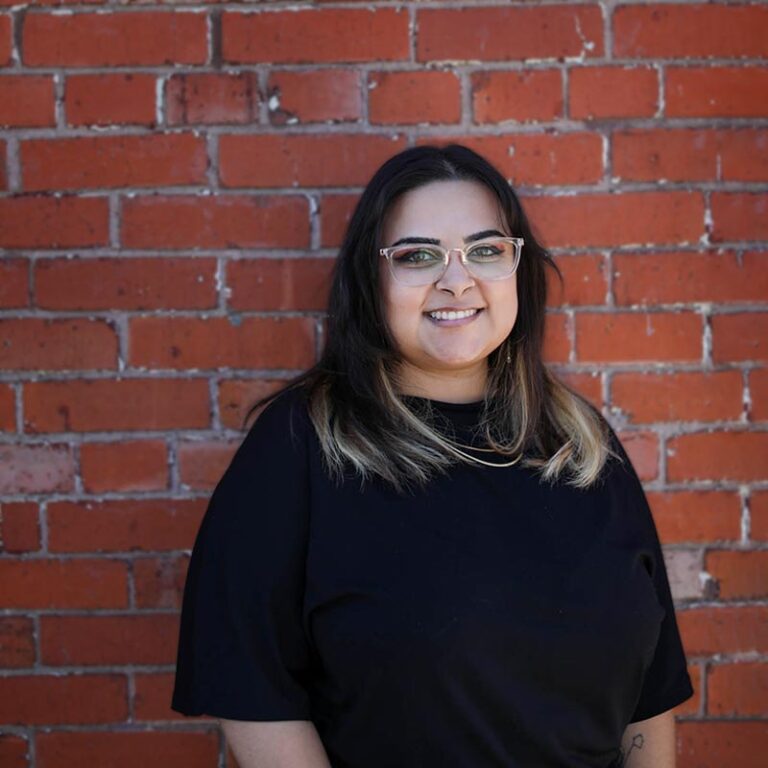 Woman with glasses smiles against a brick wall