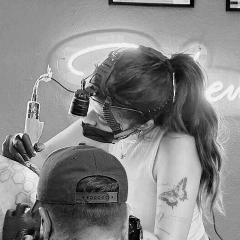 Tattoo artist with magnifying loupes applies a butterfly tattoo to a client's arm.