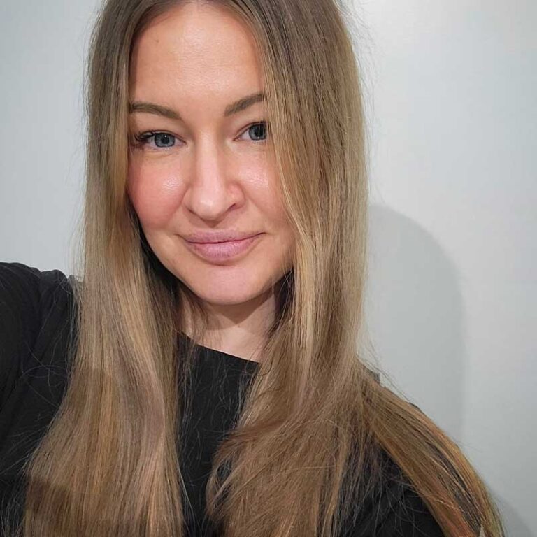 Blonde woman with blue eyes wearing black smiles at the camera with long wavy hair