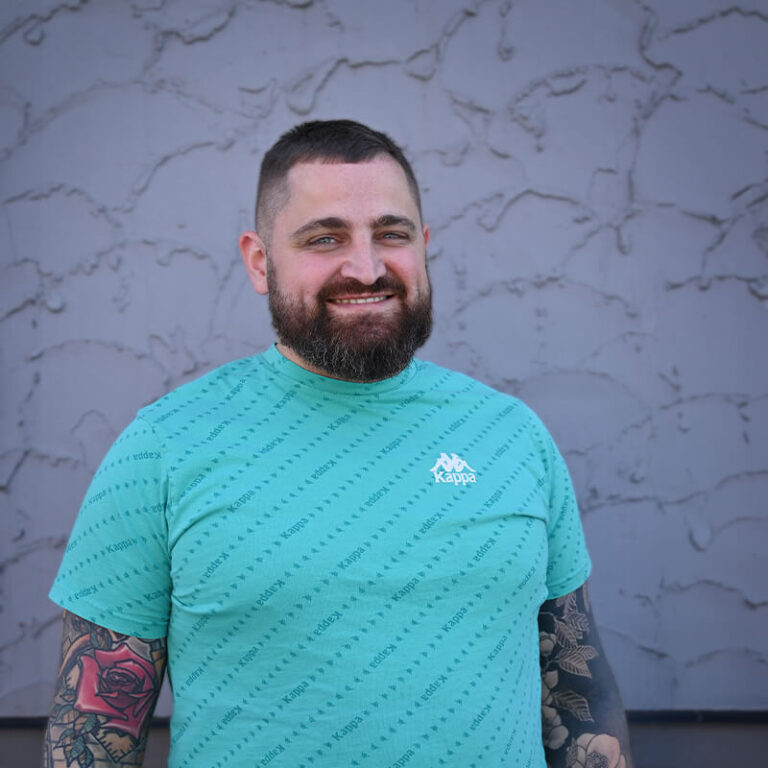 Man with beard and tattoos wearing a turquoise Kappa t-shirt with patterned logo.