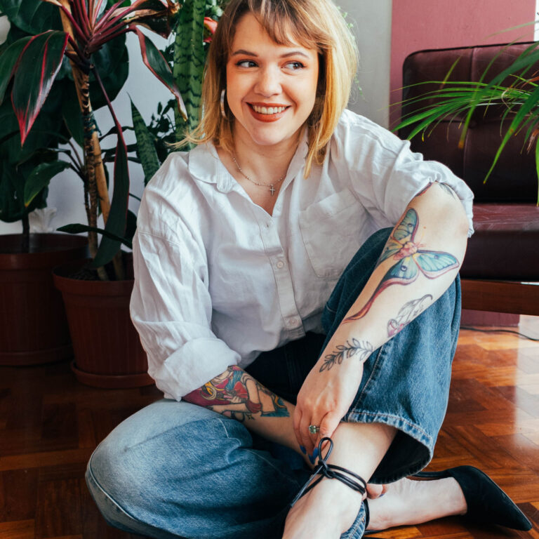 Young woman with colourful arm tattoos wearing jeans and a white shirt sits on a wooden floor surrounded by plants.