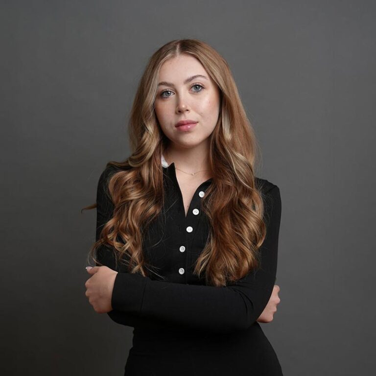 Young woman with blonde hair wearing a black shirt with white buttons on a grey background.