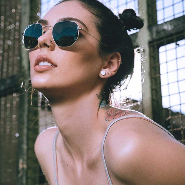 Woman in sunglasses with nose and ear piercings, flower tattoo on shoulder.
